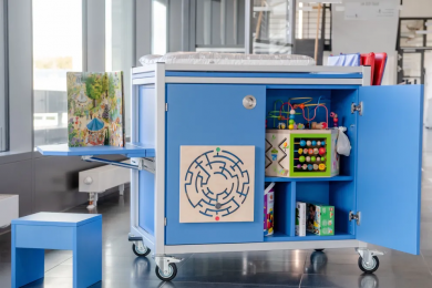 Zu sehen ist eine blaue Kommode auf Rollen, in der Spiele, Bücher gelagert sind sowie eine Wickelfläche auf der Kommode.